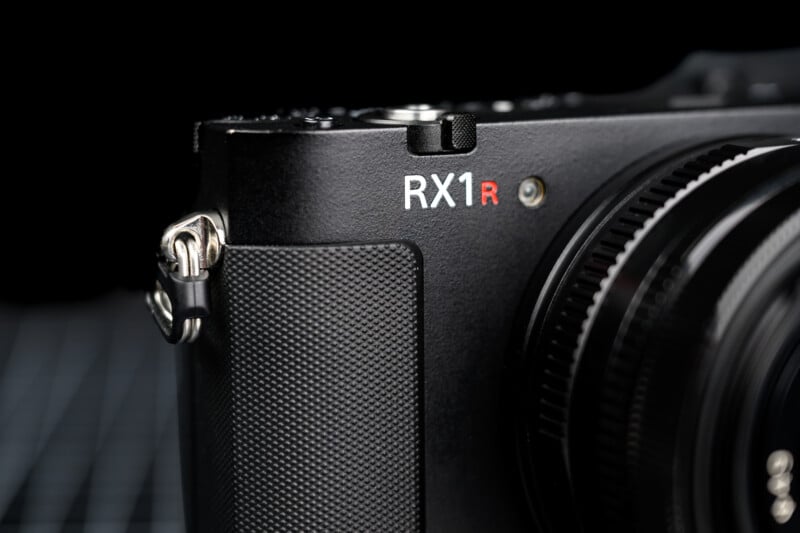 Close-up of a black Sony RX1R camera showing the textured grip, lens barrel, and part of the RX1R logo in white and red on the camera body, against a dark blurred background.