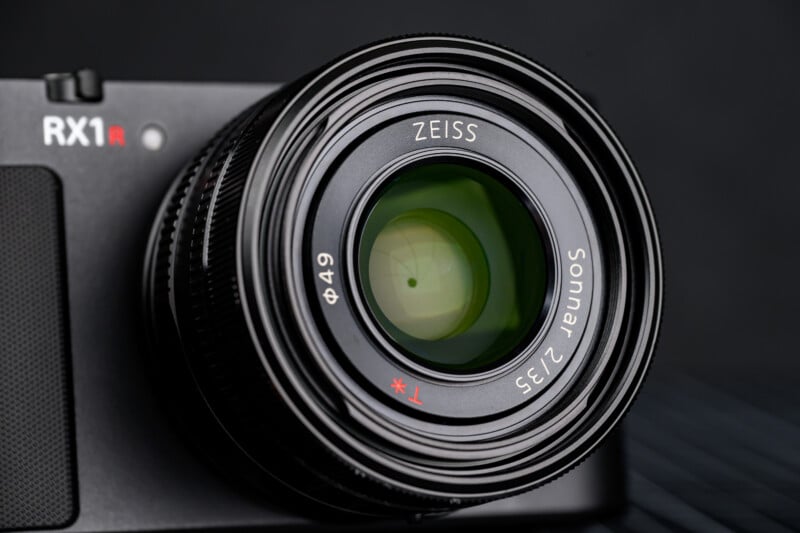 Close-up of a camera lens labeled "ZEISS Sonnar 2/35" attached to a black Sony RX1R camera body, with the lens reflecting light and the background softly blurred.