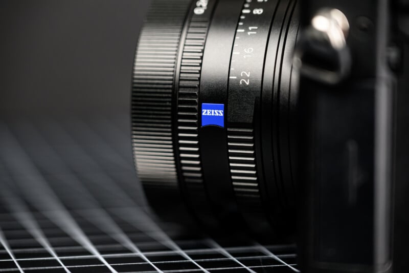 Close-up of a camera lens with a visible blue "Zeiss" logo, featuring focus and aperture markings, set against a black background with a grid pattern.