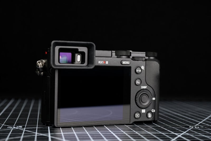 A digital camera shown from the back, featuring a large LCD screen, control buttons, and an electronic viewfinder, placed on a gridded surface with a black background.
