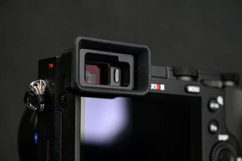 Close-up of a digital camera, focusing on the rectangular viewfinder and part of the rear screen, with a black background.