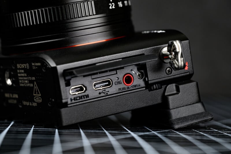 Close-up of a digital camera’s side panel showing HDMI, USB, and charging ports, labeled for connectivity and power, with a metal strap lug and part of the lens visible. The camera is resting on a grid-patterned surface.