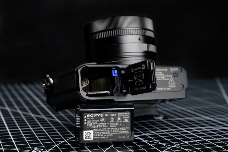 A close-up of a Sony digital camera with its battery compartment open, showing the NP-FW50 battery removed and positioned in front of the camera on a black grid-patterned surface.