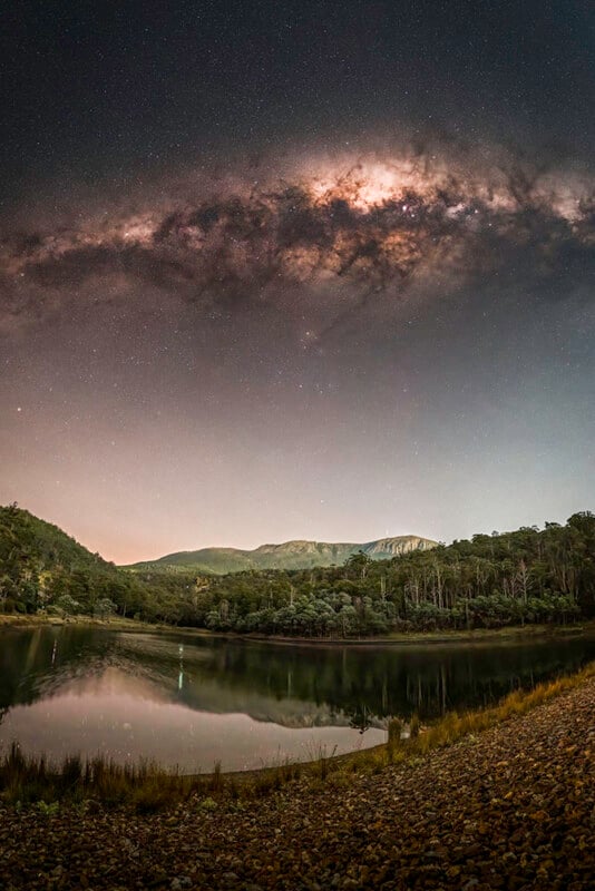 The Milky Way stretches brightly across a starry night sky above a calm lake, surrounded by trees and distant mountains, with the scene reflected in the water.