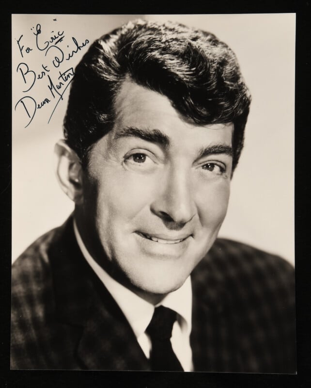 Black-and-white portrait of a smiling man with dark, wavy hair wearing a plaid jacket. There is a handwritten autograph in the upper left corner.