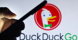 A hand holding a smartphone is silhouetted in front of a screen displaying the DuckDuckGo logo, featuring a white duck with a green bow tie inside a red circle.