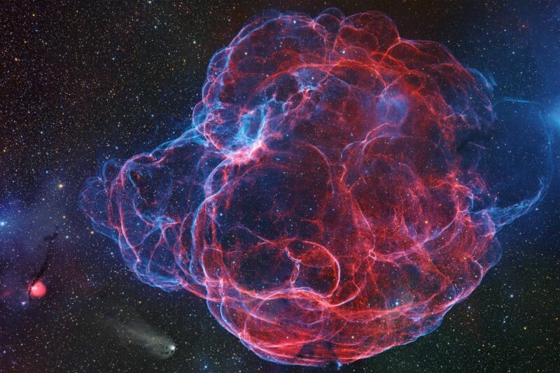 A vibrant nebula in space with wispy, glowing red and blue filaments forming a cloud-like structure against a background of stars and faint objects.