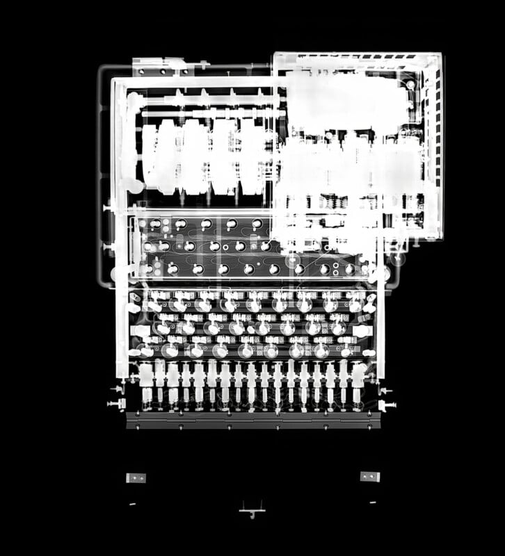 X-ray view of a complex mechanical device, possibly a vintage calculator or machine, revealing intricate inner gears, dials, and electrical components arranged in a rectangular layout against a black background.