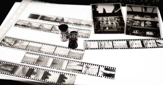 Black and white photo negatives, film canisters, and two printed photographs are displayed on a lightbox, showcasing various urban and landscape scenes.