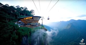 A modern, glass-walled building sits atop a lush, misty mountain cliff, with cable cars traveling over a deep valley surrounded by green mountains under a bright sky.