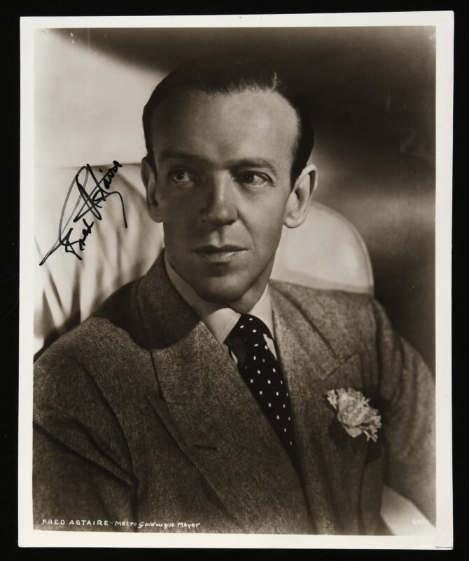 Black and white portrait of a man in a suit and polka dot tie, with a flower in his lapel, seated and looking to the side. The photograph is signed "Fred Astaire" in the top left corner.