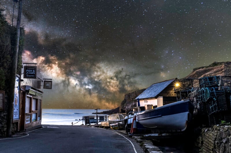 A narrow coastal road curves past a small boat shed and fishing nets, with boats on the side. The night sky above sparkles with stars and the bright arc of the Milky Way, creating a dramatic contrast over the calm sea.