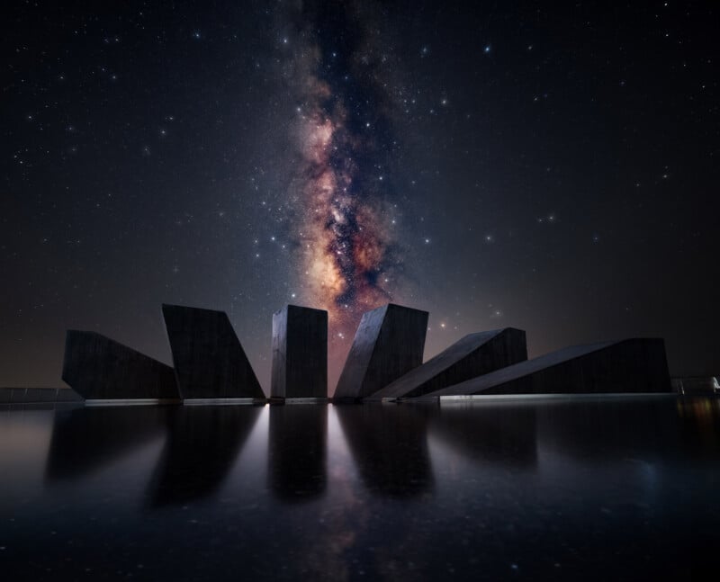 A dramatic night sky filled with stars and the glowing Milky Way above angular, dark stone structures reflected in calm water, creating a surreal and striking scene.