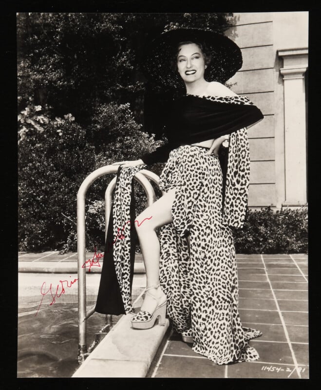 A smiling woman in a long, off-the-shoulder leopard print dress and large black hat poses by a swimming pool ladder, one leg raised to reveal a high slit, with a towel draped over the rail.
