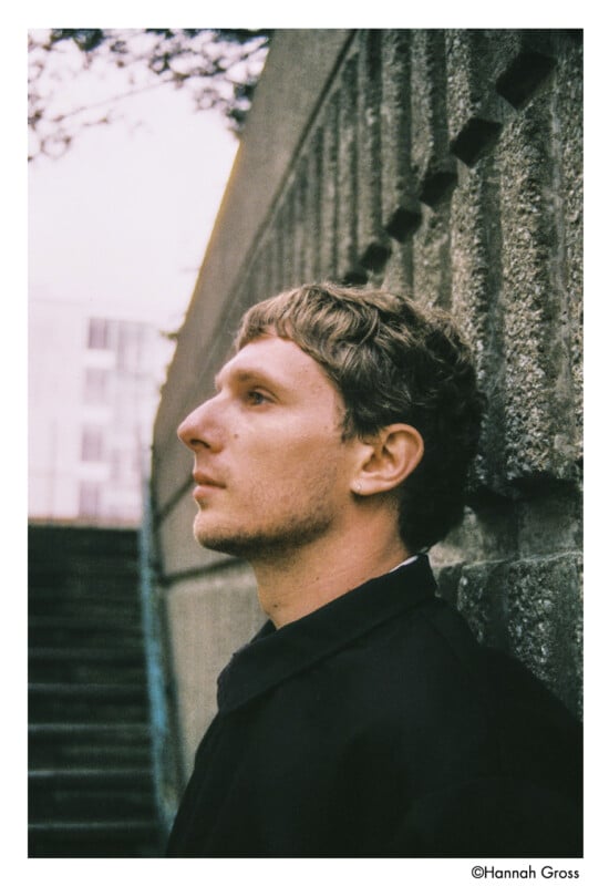 A person with short, light brown hair stands in profile next to a concrete wall and stairs, looking contemplative. The image has a soft, muted tone and an outdoor, urban setting.