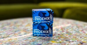 Two boxes of Harman Phoenix ISO 200 colour film stacked on a colorful, speckled surface with a blurred green background. The film boxes are blue with bold, stylized graphics and text.