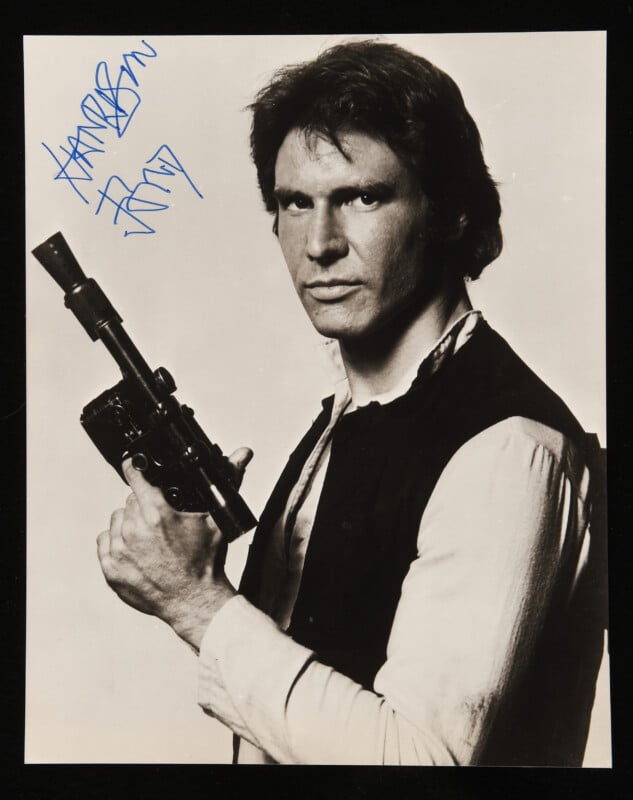 A man with medium-length hair, wearing a vest over a light shirt, holds a sci-fi blaster. He looks seriously at the camera. The image is in black and white and features a blue signature in the top left corner.