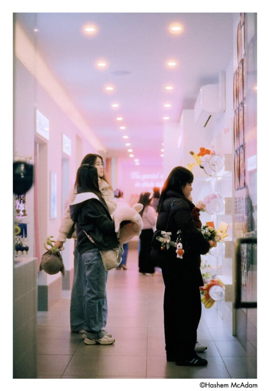 Two young women stand in a softly lit hallway; one holds a bouquet and a plush toy, while the other looks at posters on the wall. More people are further down the hallway, which glows with pink and white lights.