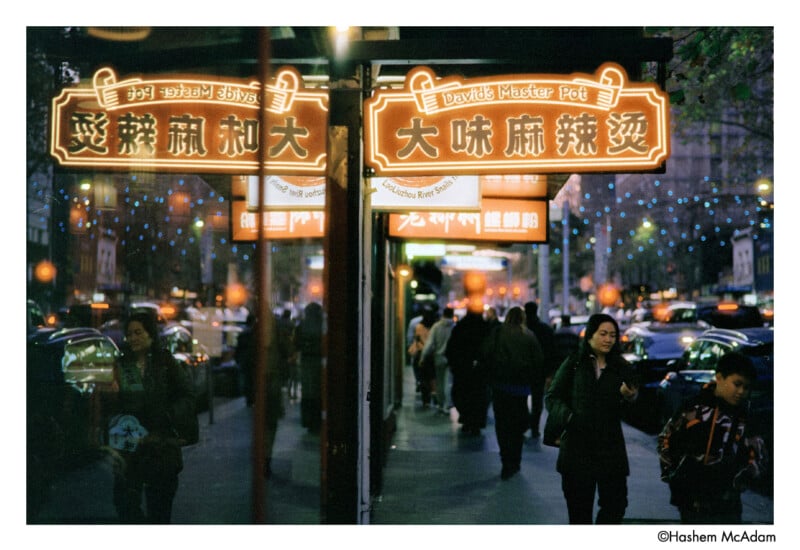 A busy city street at night with glowing Chinese restaurant signs, people walking by, and reflections of lights and signs in the shop windows.