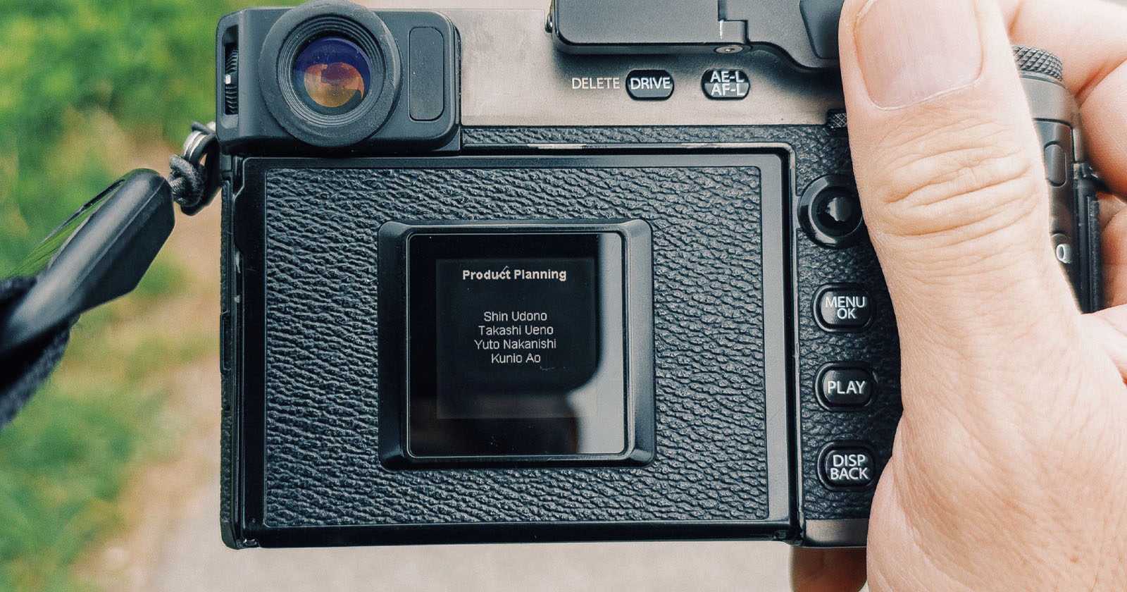 A close-up of a person holding a digital camera, showing its screen displaying a “Product Planning” credits list with four names. The camera’s buttons and textured grip are visible, with a blurred outdoor background.