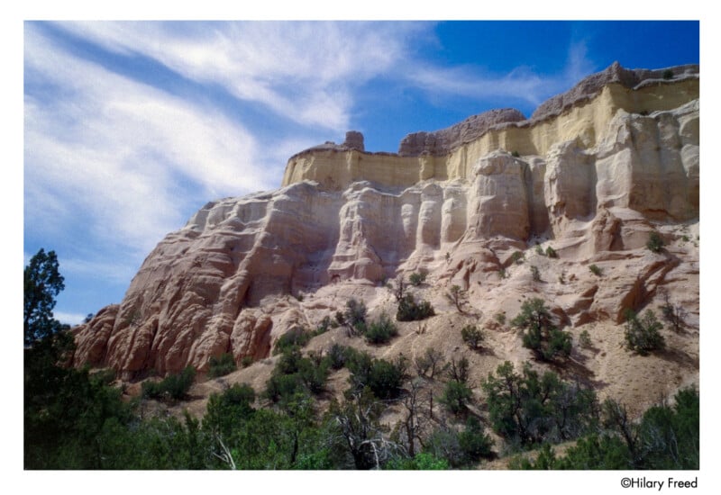 Layered, colorful sandstone cliffs rise above desert vegetation under a blue sky with wispy clouds. The rock formations show distinct bands of cream, tan, and reddish hues.