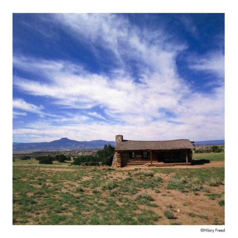A small log cabin with a stone chimney sits on a grassy prairie under a wide, partly cloudy blue sky, with distant mountains on the horizon.
