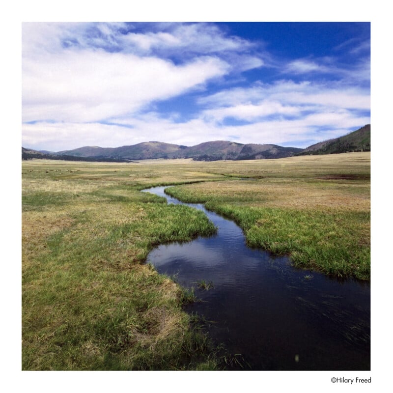 A small stream winds through a grassy meadow under a blue sky with scattered clouds, with distant hills and mountains in the background.