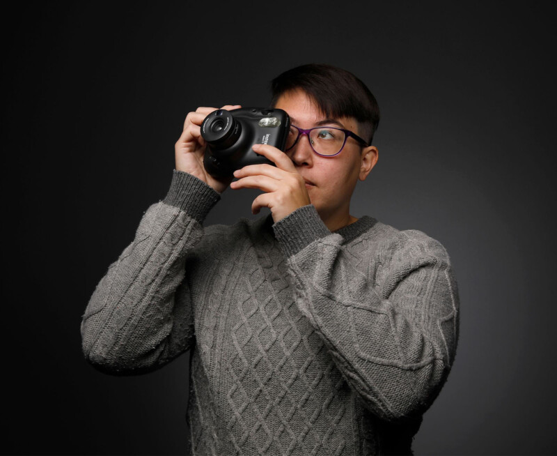 A person wearing glasses and a gray sweater holds a camera up to their face, appearing to take a photo against a dark, plain background.