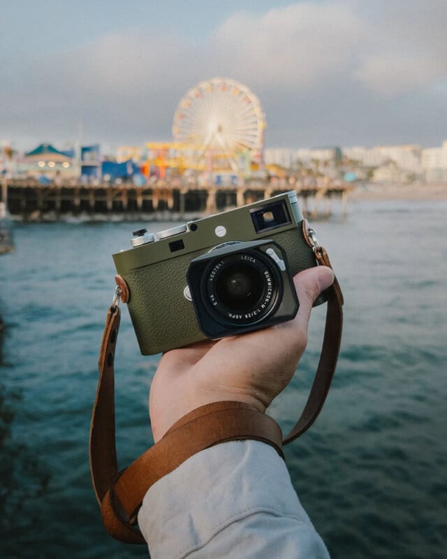 A hand holds a green vintage-style camera with a brown strap in front of a blurred background of an amusement park, featuring a Ferris wheel near the water.