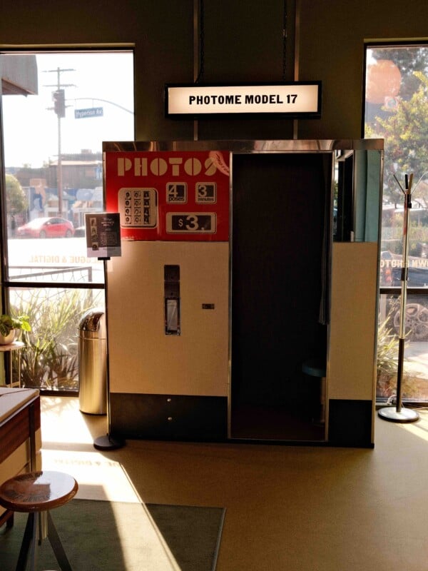 A vintage photobooth labeled "PHOTOME MODEL 17" stands in a sunlit room near large windows. Signs show photo examples and pricing, and outdoor scenery is visible through the glass.