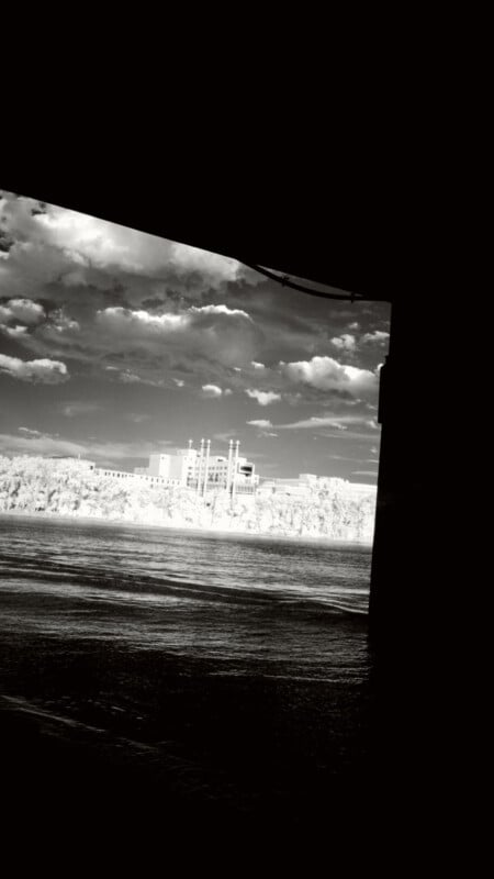 A black-and-white photo taken from under a bridge, looking out over a wide river toward a building surrounded by trees, with a cloudy sky overhead.