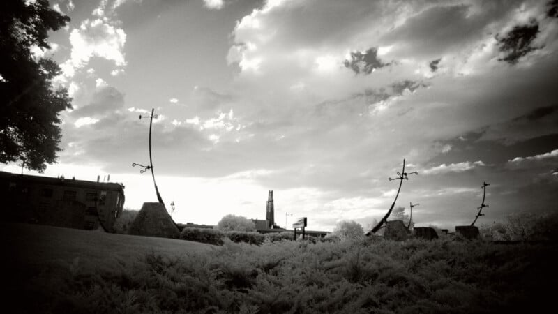 A black and white photo of a park with curved metal sculptures, bushy plants in the foreground, and dramatic clouds in the sky. Buildings and a tall tower are visible in the background.