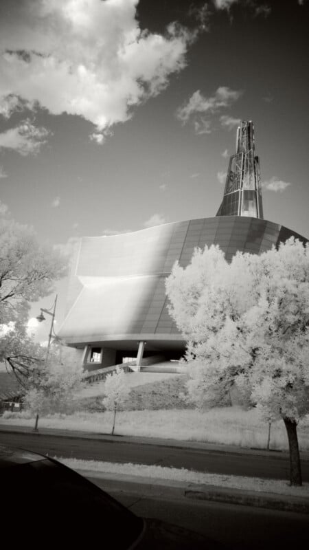 A modern, angular building with a tall glass spire rises behind leafy trees, photographed in black and white. The sky is partly cloudy and a parked car is visible in the foreground.
