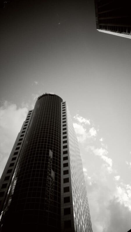Black and white photo of a tall, modern skyscraper with a cylindrical center and rectangular windows, viewed from below against a partly cloudy sky. Another building is partially visible in the top right corner.