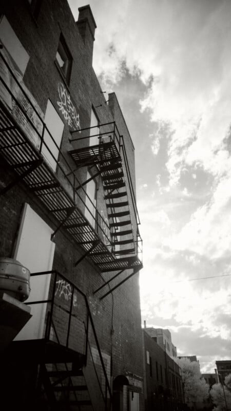 Black and white photo of a brick building with graffiti, featuring metal fire escapes zigzagging up the side. The sky is partly cloudy, and the scene has an urban, industrial atmosphere.