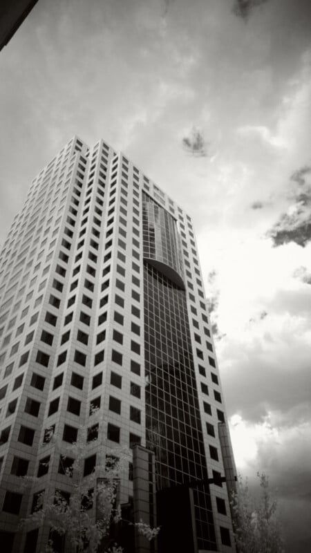 Black and white photo of a tall, modern office building with a grid-like pattern of windows, set against a cloudy sky. The angle is from below, emphasizing the building’s height and geometric design.