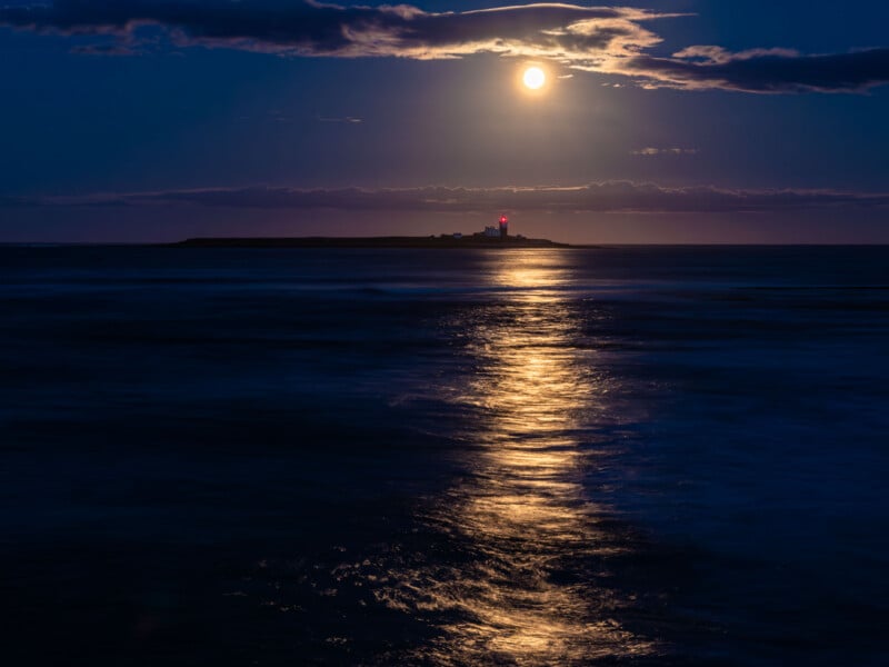 A bright full moon illuminates the night sky and reflects on the calm sea, with a distant island and a lighthouse with a red beacon visible on the horizon.