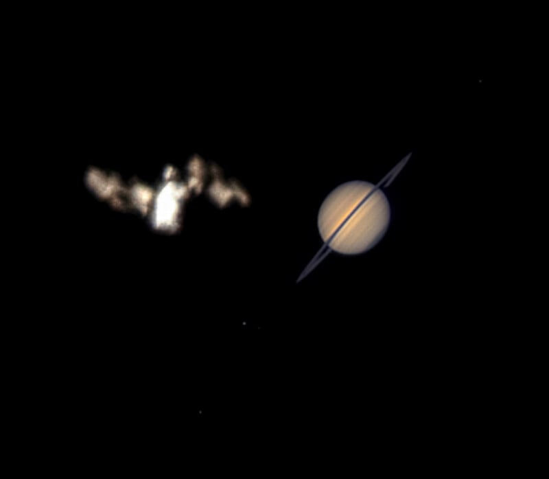 A bright, cloudy object appears near Saturn, which is shown with its rings against a black background of space. Saturn is clearly visible, while the other object is blurry and irregularly shaped.