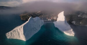 Aerial view of two large icebergs floating in blue-green water near a coastal village with mist and rocky terrain in the background.