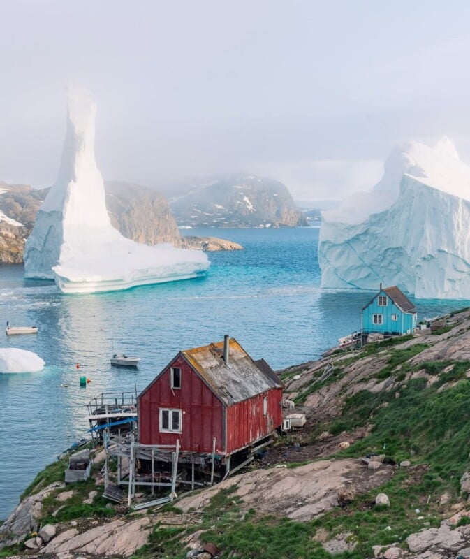 A red wooden house and a blue house sit on a rocky slope overlooking calm water with large icebergs floating nearby, under a cloudy sky in a remote Arctic coastal landscape.