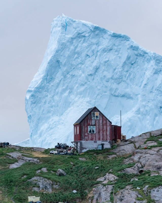 A small, weathered red house stands on a rocky hillside with scattered debris, while a massive blue iceberg towers in the background under an overcast sky.