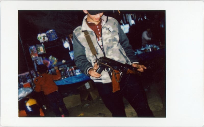 A person in a patterned jacket and cap holds a toy gun at an outdoor market stall, with colorful items and another child blurred in the background.