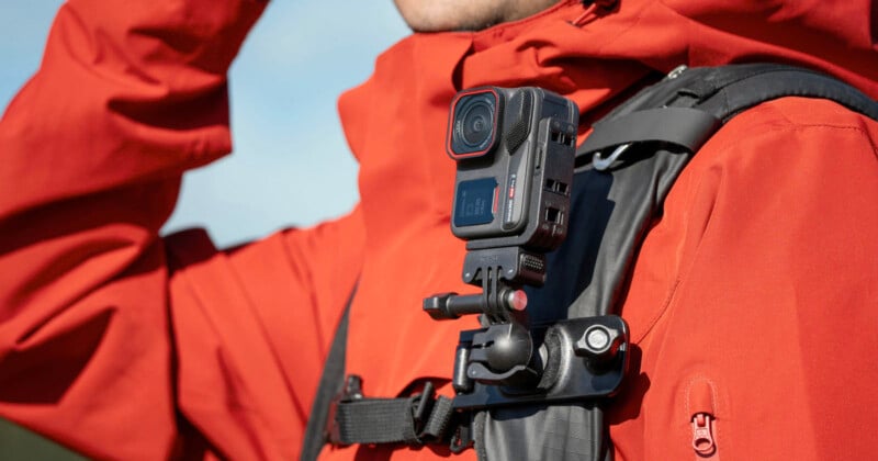 A person wearing a red jacket has a black action camera mounted on the strap of their black backpack, ready for hands-free recording outdoors.