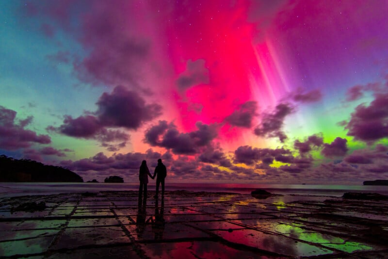 Two people stand holding hands on a rocky shoreline, silhouetted against a vibrant sky lit by colorful auroras in shades of pink, purple, green, and blue, with dramatic clouds and stars above.