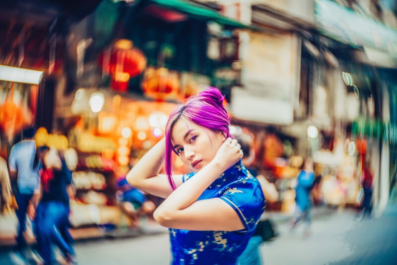 A person with vibrant purple hair, wearing a blue patterned dress, poses confidently on a bustling street with colorful lights and blurred people in the background.