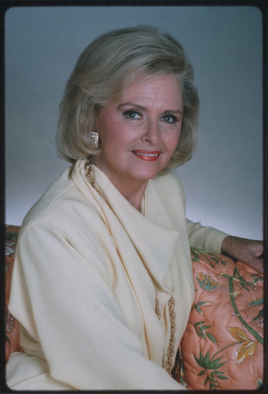 A mature woman with short blonde hair and pearl earrings, wearing a cream-colored outfit and gold chain necklace, sits on a peach floral-patterned couch, smiling at the camera against a plain background.