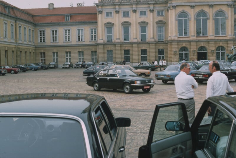 A cobblestone courtyard filled with parked cars outside a large historic building; two men in white shirts stand in the foreground near an open car door, while another car drives through the lot.