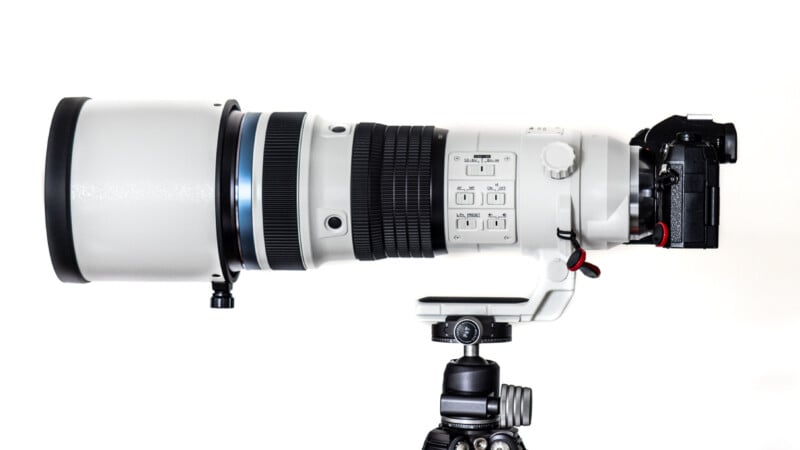 A large white telephoto camera lens mounted on a black tripod head, attached to a digital camera, set against a plain white background.