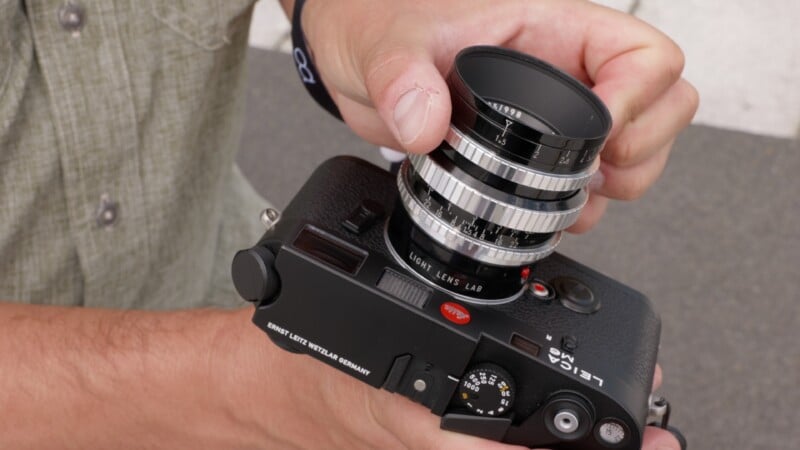 A person’s hands attach a silver camera lens to a black Leica camera body, with part of their green shirt and a wristband visible in the background.