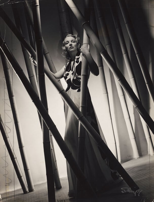 A woman in a long, patterned dress stands among large bamboo poles in a black and white photo, looking upward with a dramatic expression. Light and shadows create an atmospheric, artistic scene.